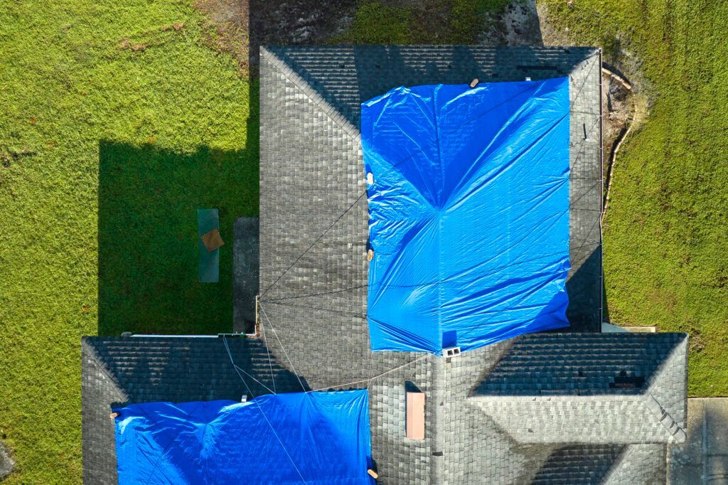 Aerial view of rooftops covered in blue tarps beside a grassy yard and paved driveway.