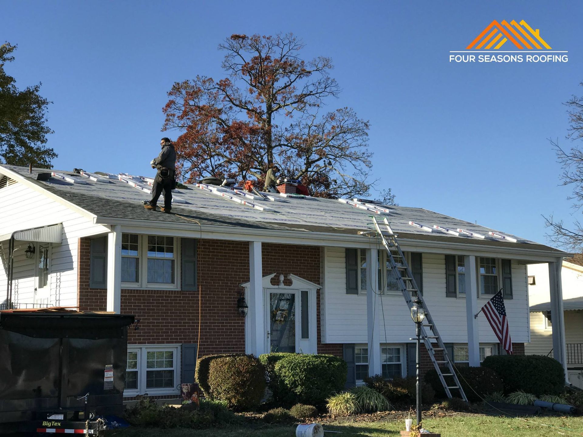 Contractors replace the shingles on the roof of a two-story brick and white house on a sunny day.