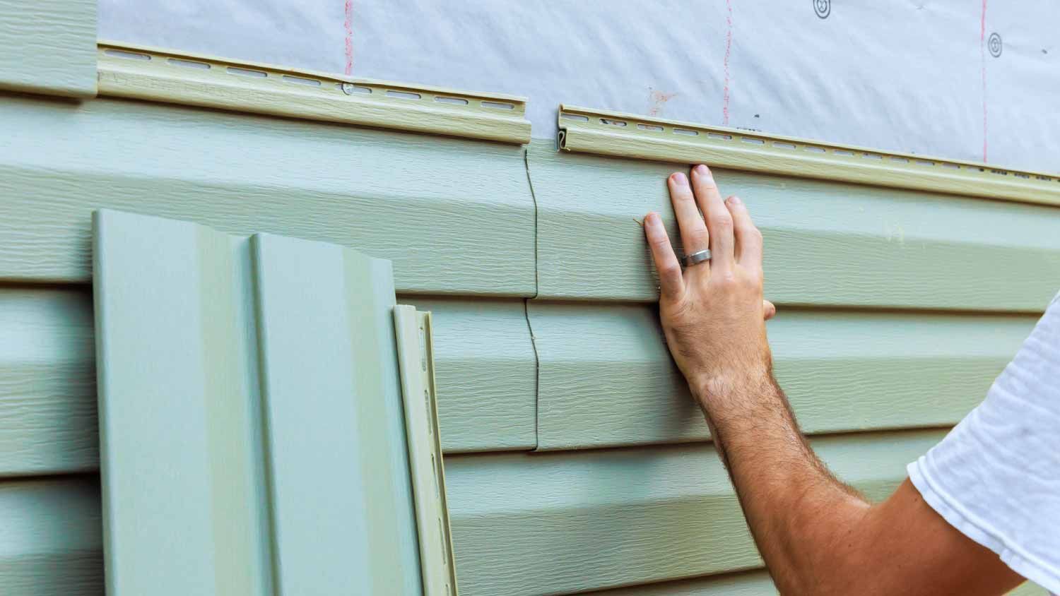 A person installing light green vinyl siding panels onto the exterior wall of a building.