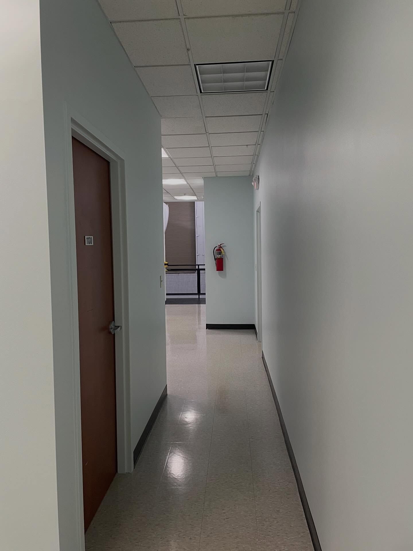 Narrow, light-colored hallway with doors and a fire extinguisher. Ceiling lights illuminate the space.