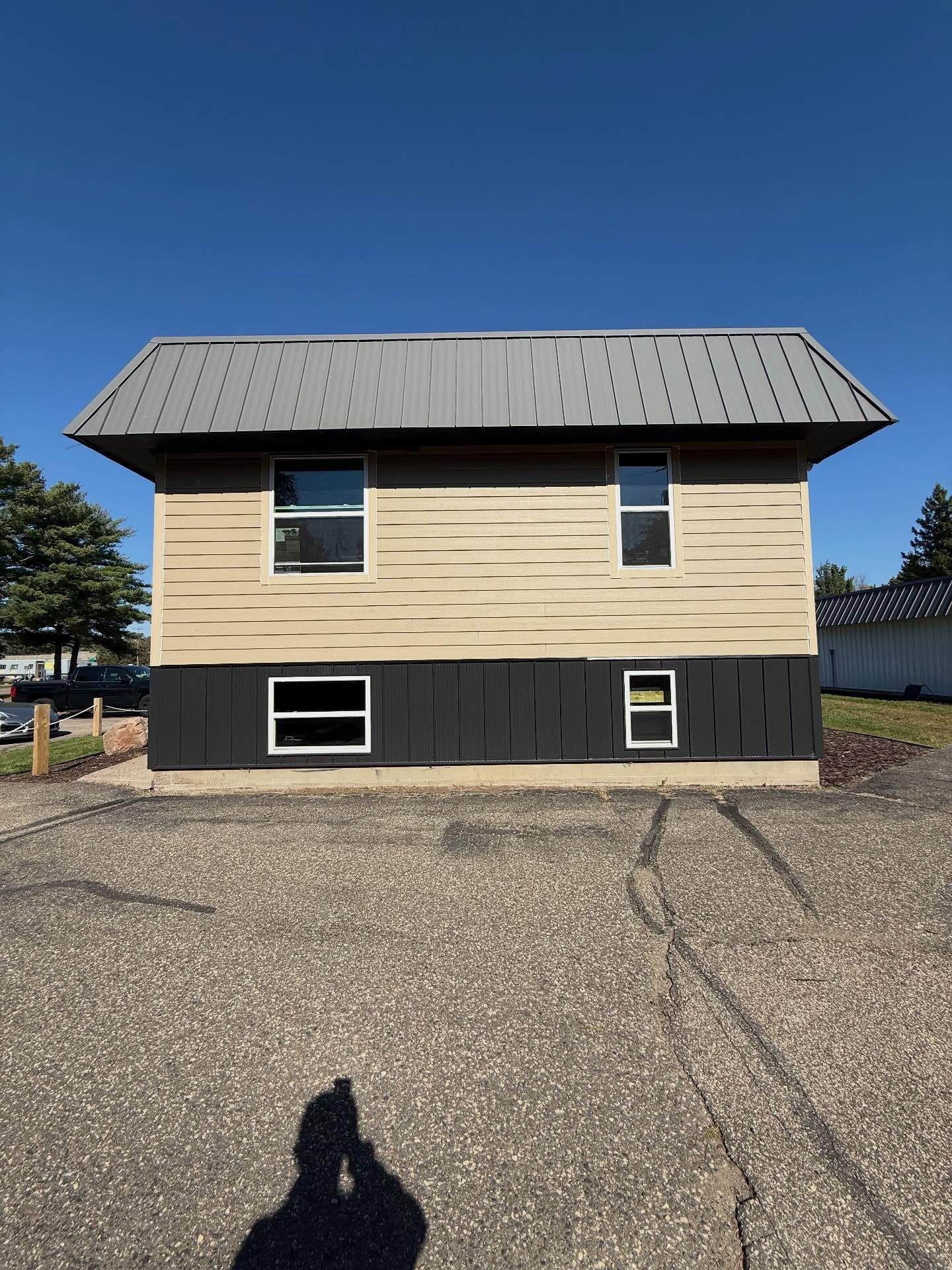 Two-story building with gray roof, black and beige siding, four windows, and a gravel parking lot.