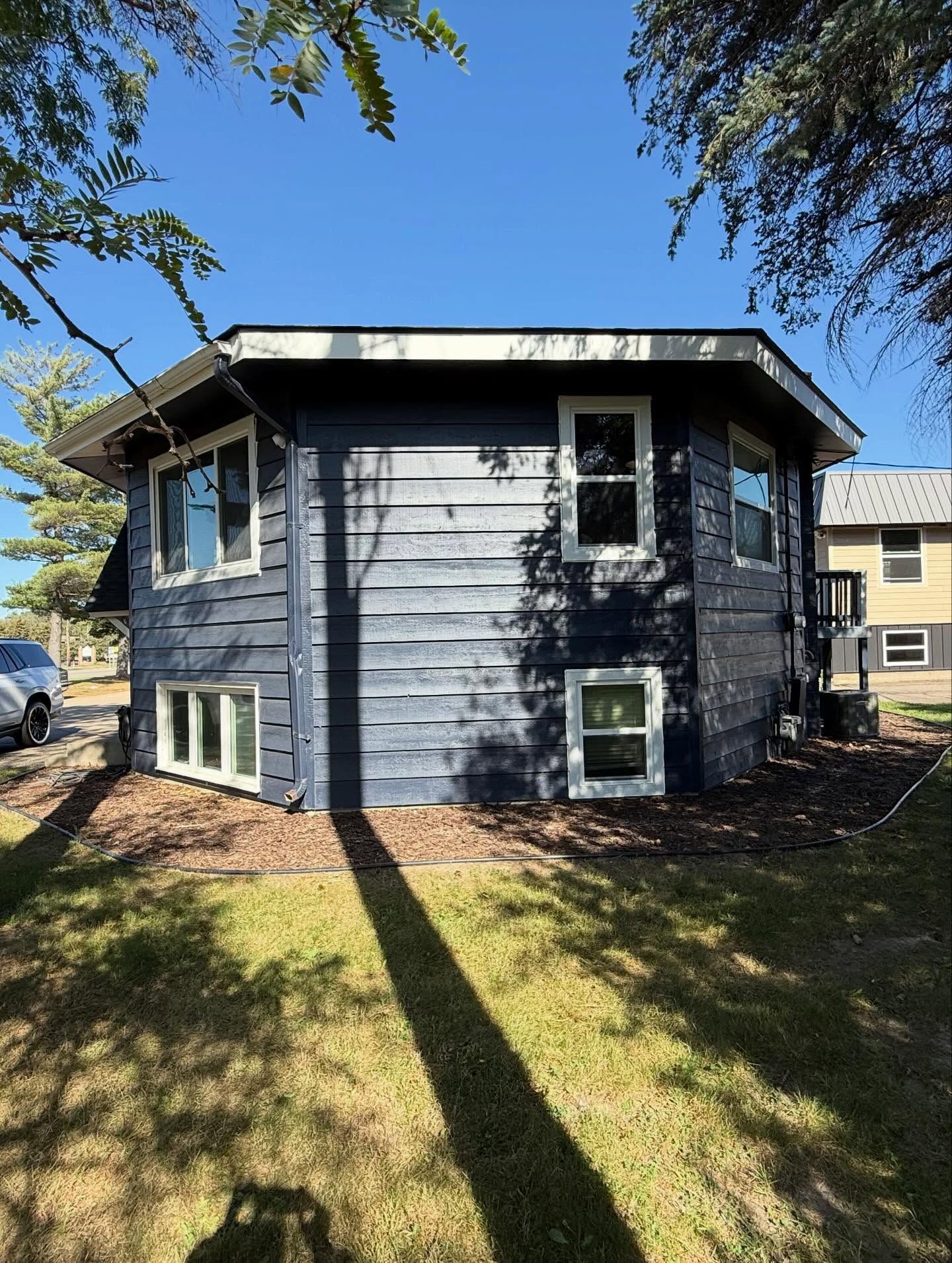 Two-story blue house with white-trimmed windows; lawn in front, trees on the side, sunny day.