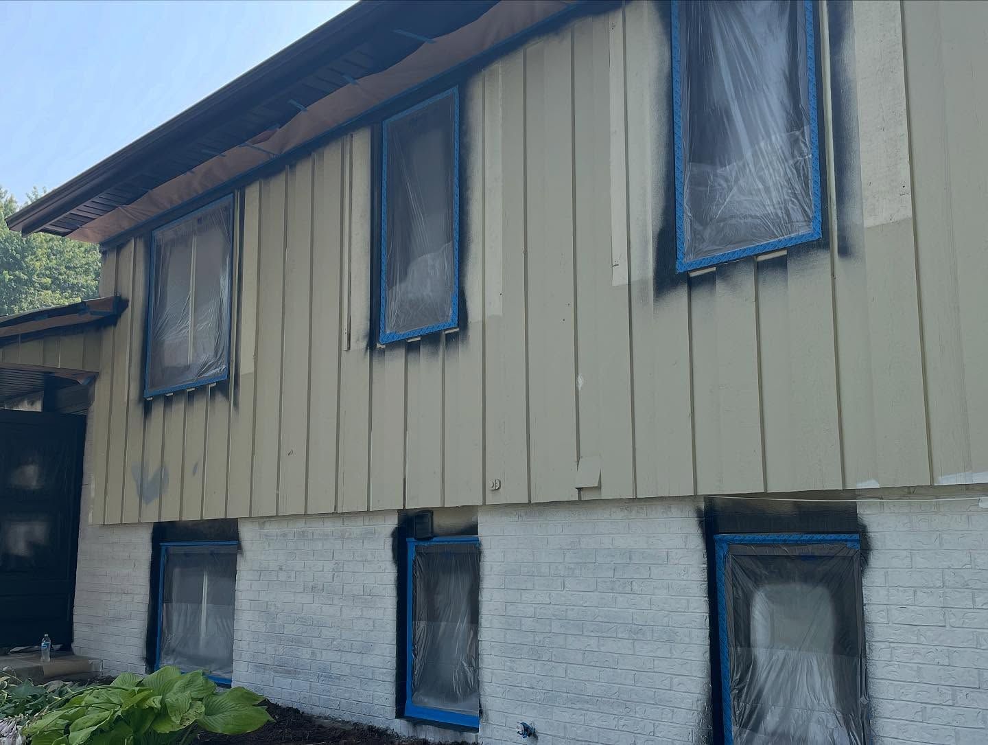 House exterior being painted; siding above a white brick facade. Windows covered in plastic. The siding is light beige.