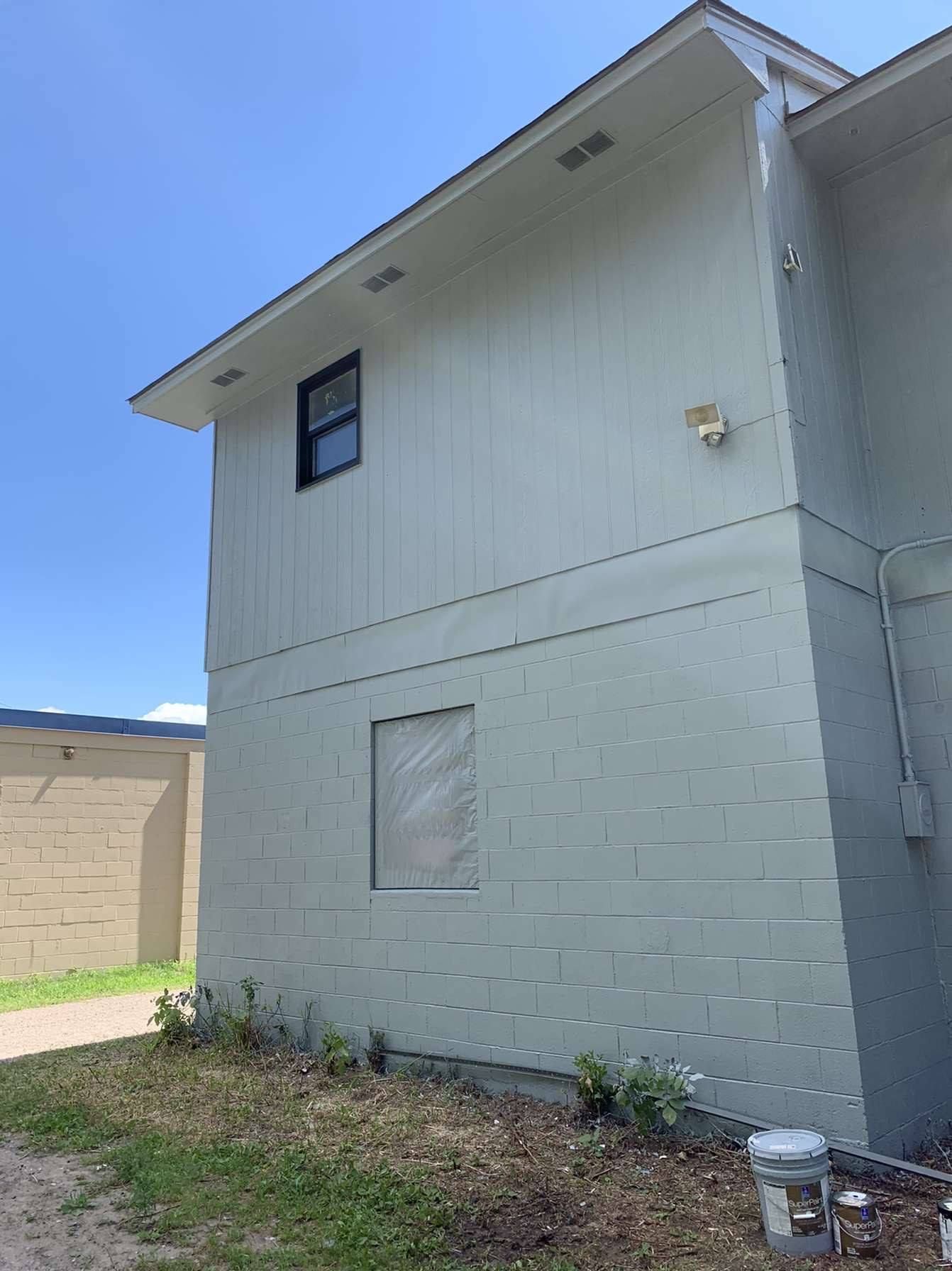 Exterior side of a two-story building with light blue siding, a small window, and a boarded-up opening. A concrete wall is to the left.