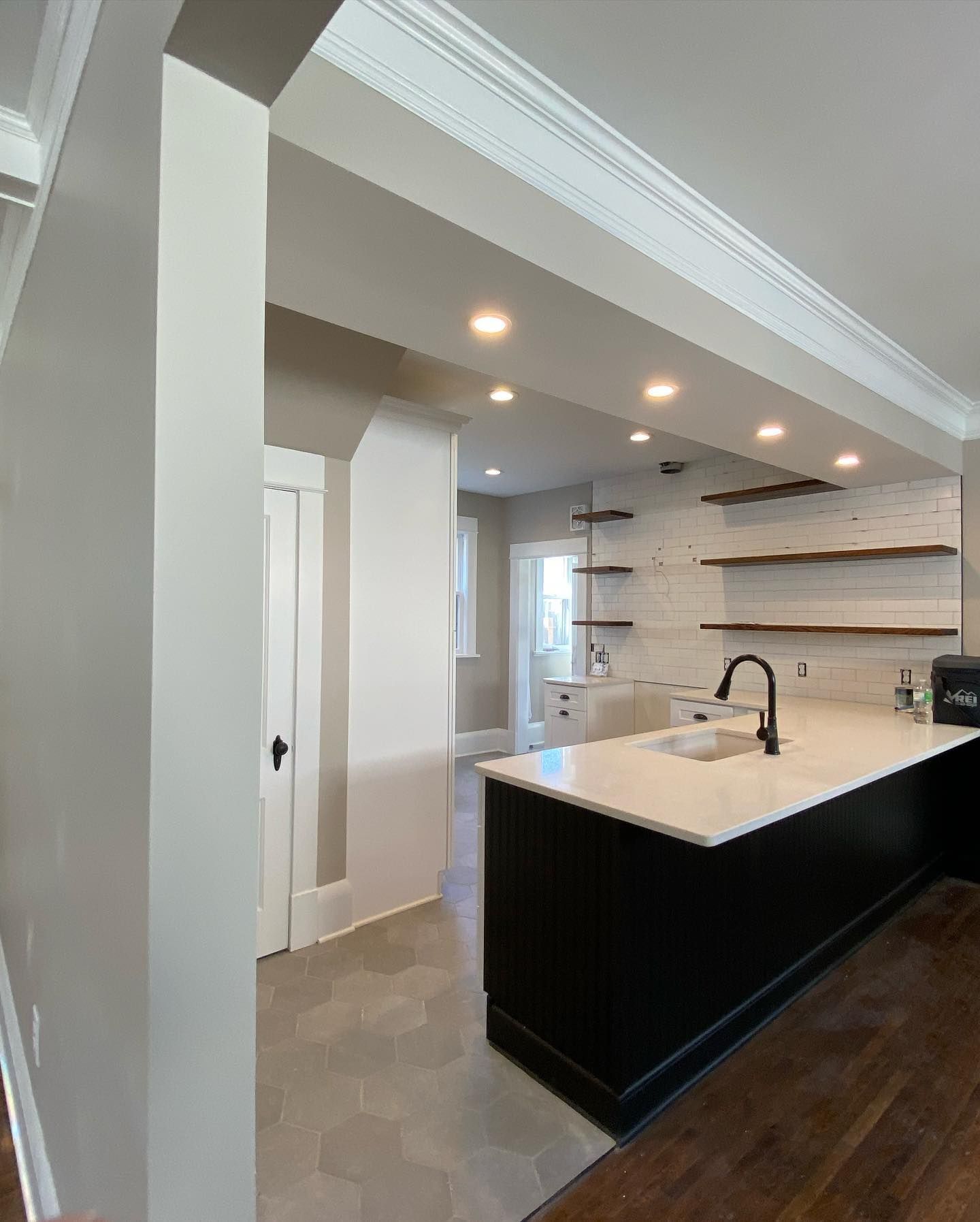 Modern kitchen with a black island, white countertops, and open shelves. A doorway leads to a hallway with a white door.