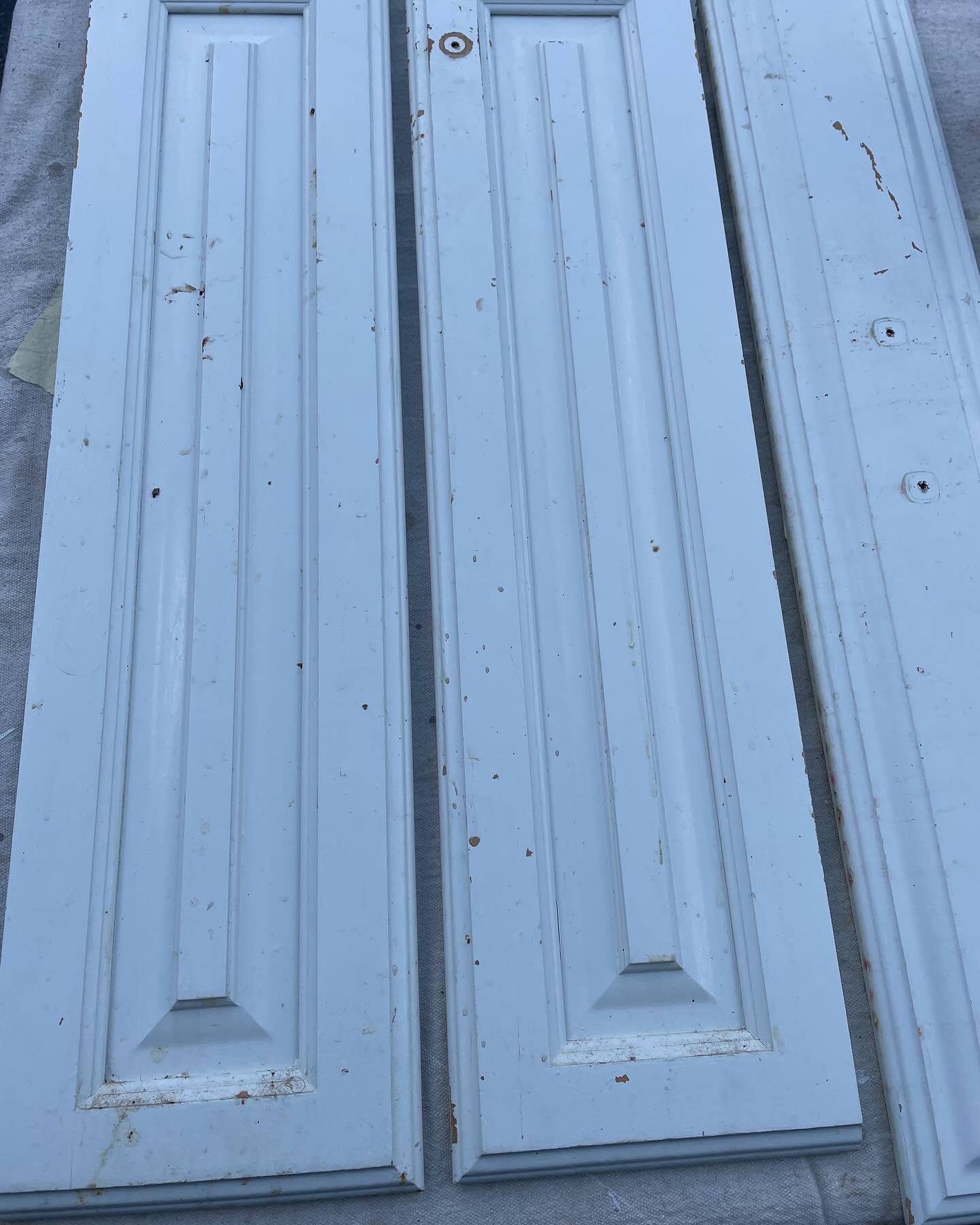 Three weathered, off-white wooden door panels laid flat. Panels have vertical grooves and rectangular accents, showing signs of paint peeling.