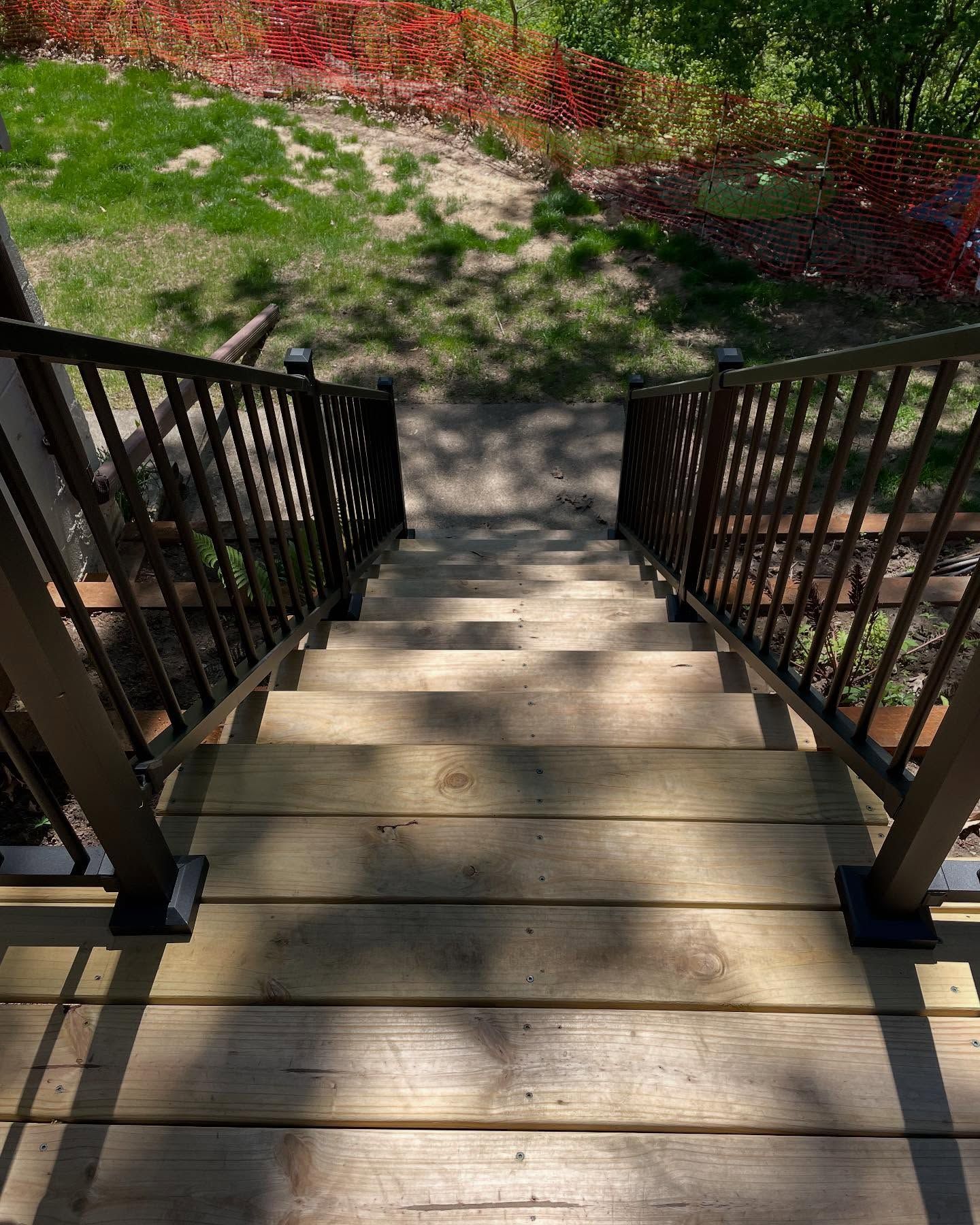 Wooden outdoor stairs with dark metal railings descend towards a grassy area with red foliage visible in the background.