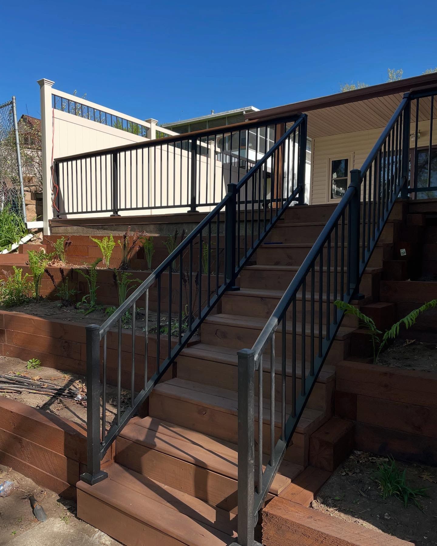 Wooden steps leading up to a house with black railings. The house has a deck with cream-colored railing.