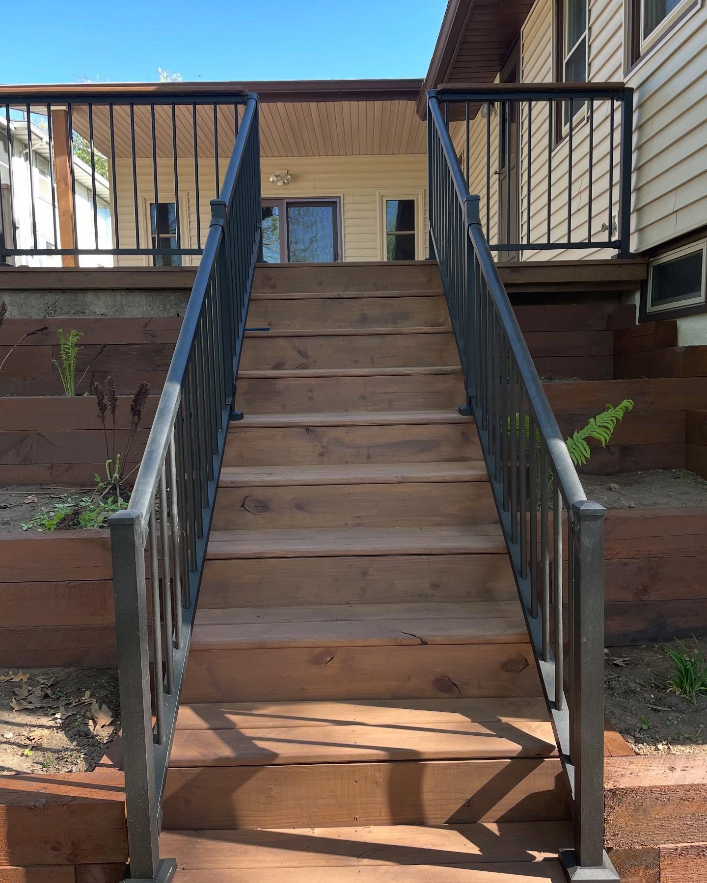 Wooden staircase with black metal railings leading up to a house. The stairs are flanked by wood retaining walls with greenery.