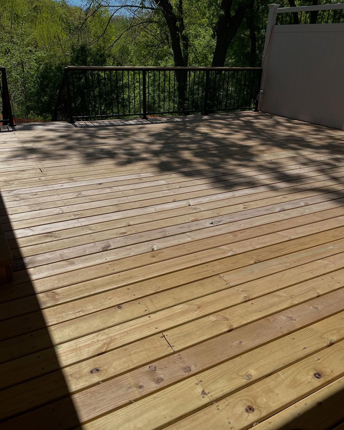 Wooden deck with sunlight and shadows, surrounded by a railing and trees. The deck appears to be empty.