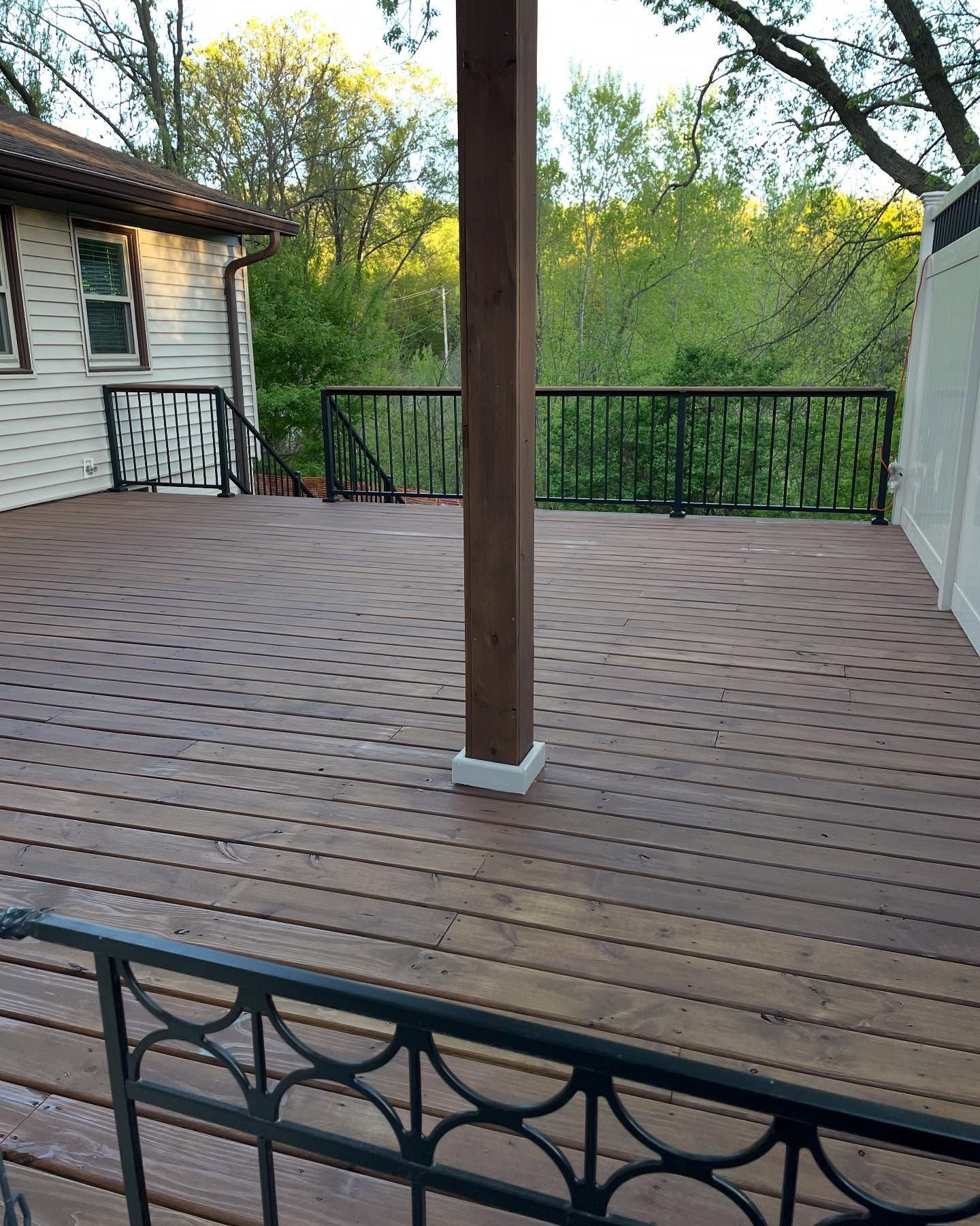 A large wooden deck with black railings and a central wooden post; the deck is adjacent to a house and overlooks greenery.