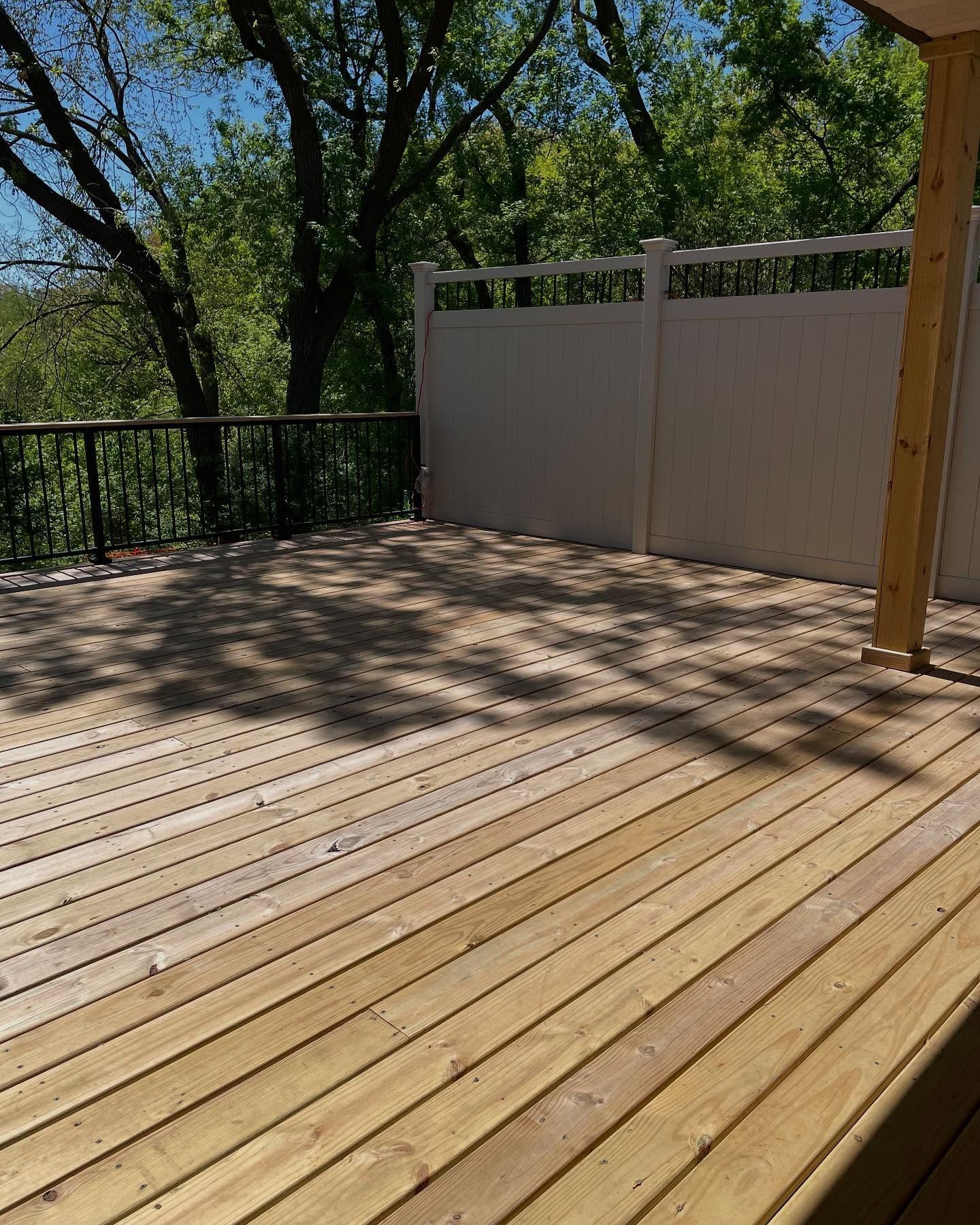 Wooden deck with a white fence and railing, surrounded by trees. Sunlight casts shadows across the deck.