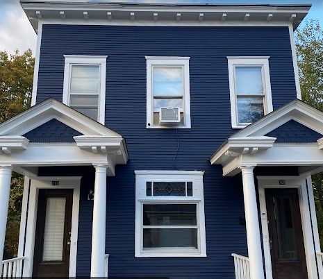 Two-story blue duplex with white trim. Each unit has a porch, front door, and multiple windows. An air conditioner is visible.