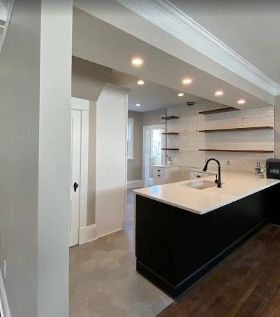 Kitchen remodel with a black island, white countertops, open shelving, and a view into an adjacent room. Gray and wood tones dominate.