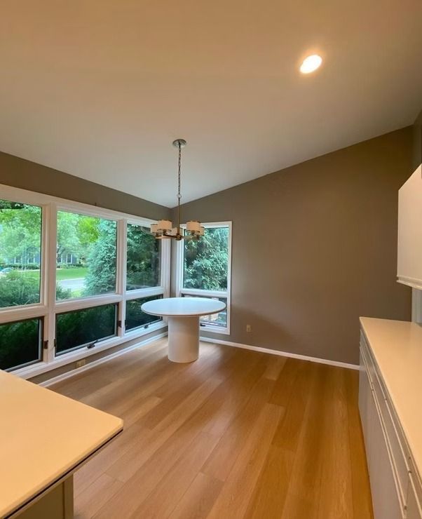 Dining area with a white pedestal table under a chandelier, hardwood floors, and large windows overlooking greenery. The walls are painted a neutral brown color.