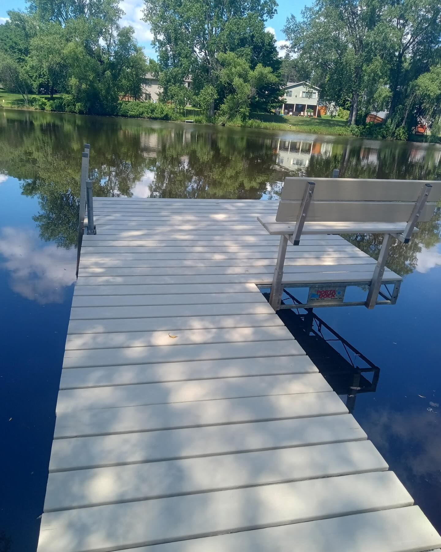 White dock with bench on a calm lake. Trees and houses are reflected in the water under a blue sky with clouds.