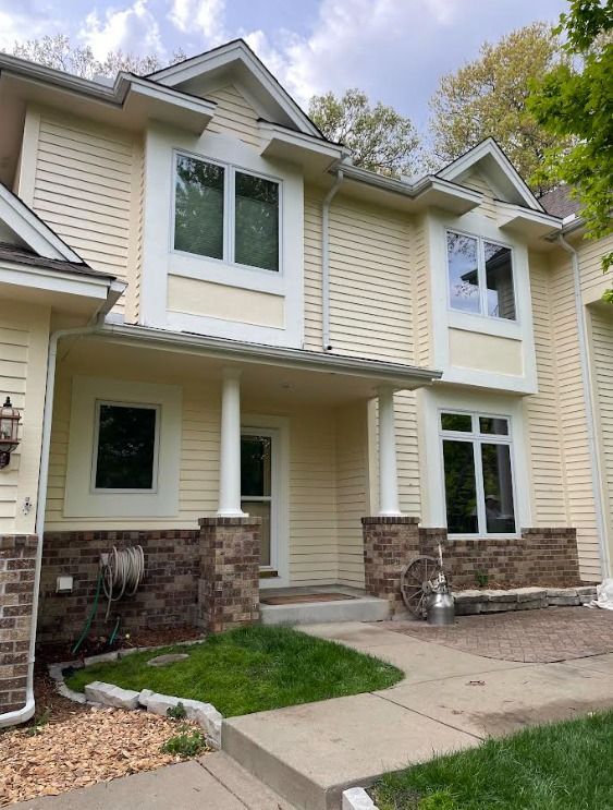 Two-story townhouse with yellow siding and white trim, featuring brown brick accents on pillars and foundation, and green lawn.