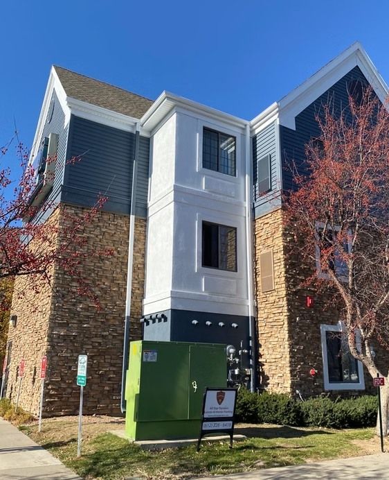 Three-story building with blue, stone, and white facades under a blue sky. A green electrical box and a sign are in front.