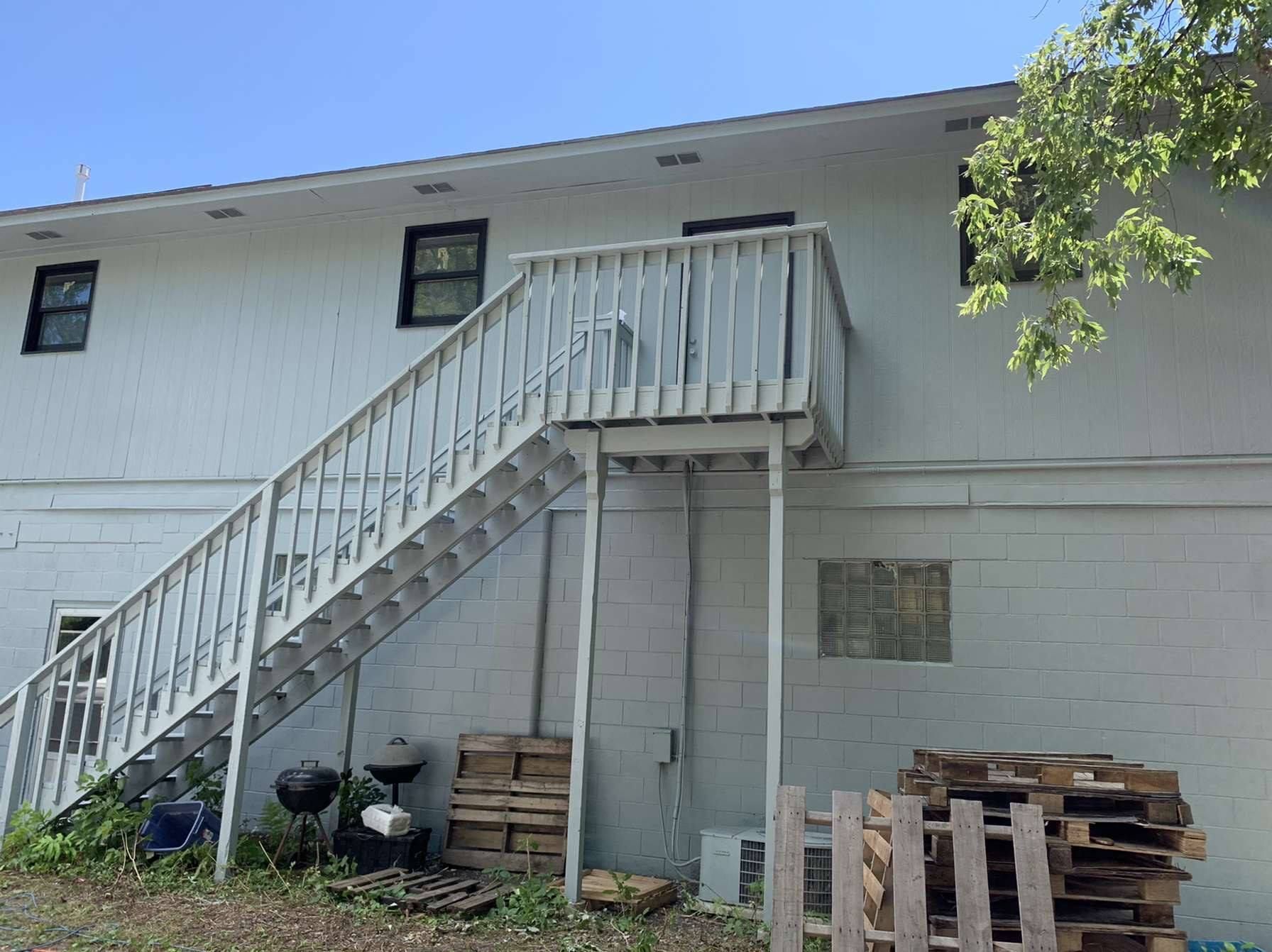 Exterior of a two-story white building with a wooden staircase leading to a second-floor deck. Pallets and a grill are below.