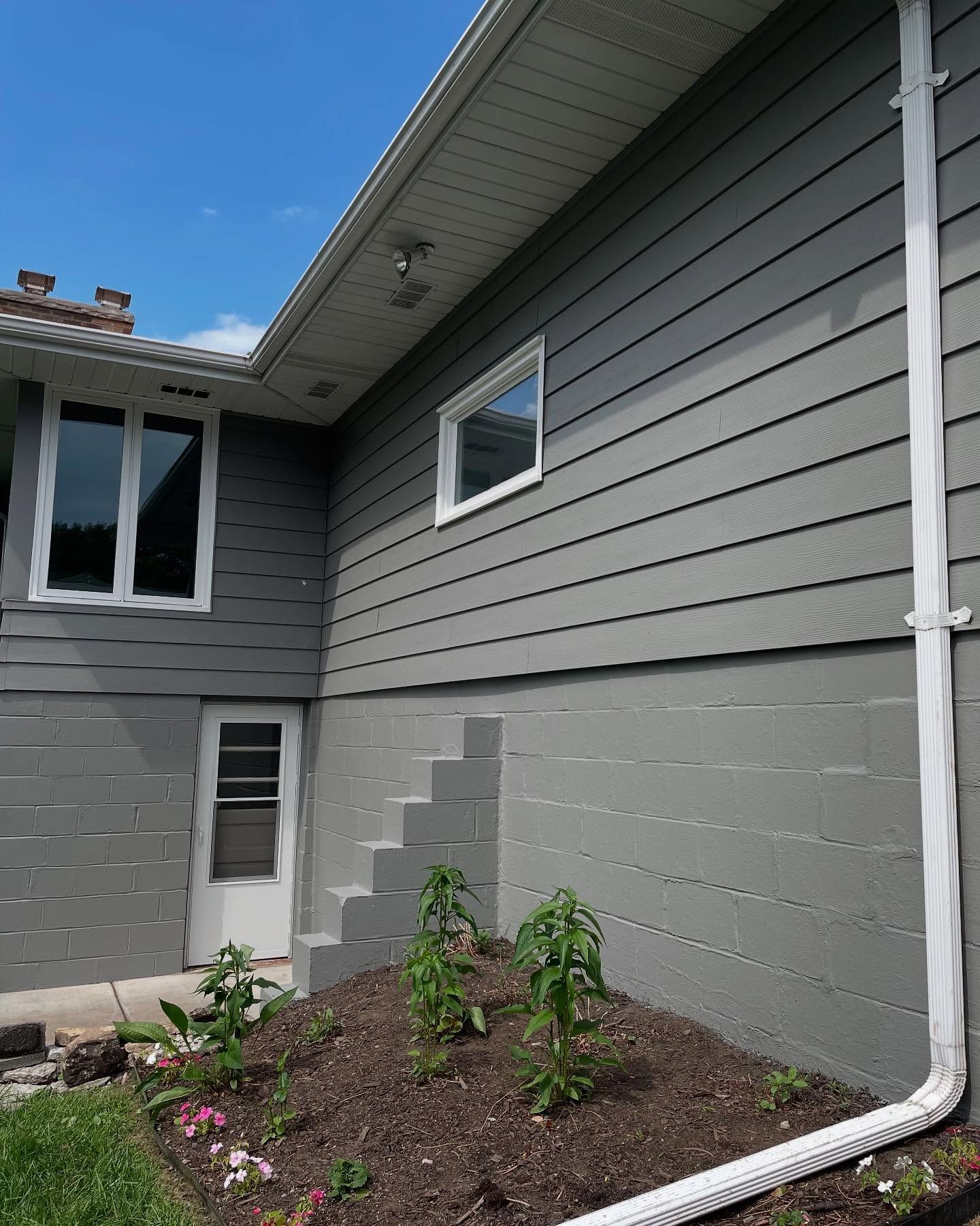 Gray house exterior with white trim and downspout, featuring a window, door, and small concrete steps in a garden bed.