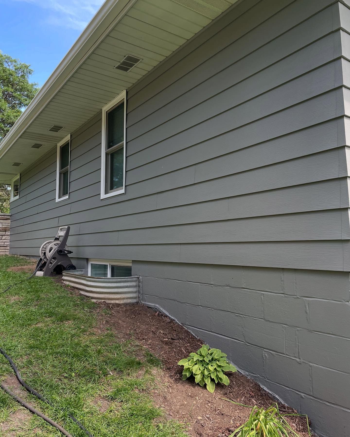Gray-sided house with white trim and two windows. A small garden bed with greenery sits below a window well, with a sloping lawn in front.