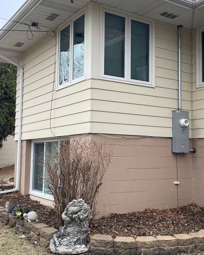Exterior of a house with beige siding, white-framed windows, and a brown concrete foundation. Electrical panel on the right side.