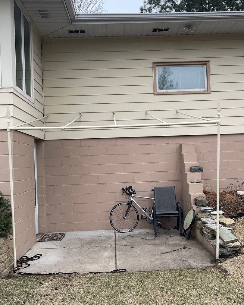 A small patio with a metal frame overhang next to a tan house. A bicycle, chair, and doormat are on the concrete.