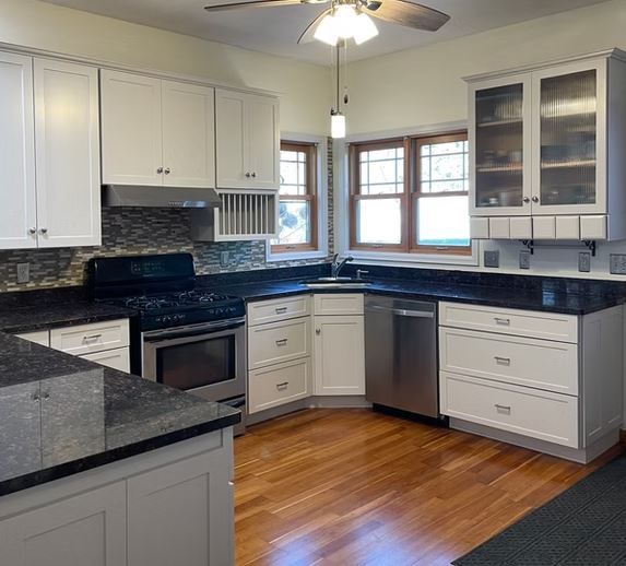 A kitchen with white cabinets, dark countertops, stainless steel appliances, and hardwood floors. There are two windows with brown frames and a glass-front cabinet.
