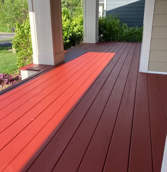 A red-painted wooden porch bathed in sunlight, with white columns and a green bush in the background.