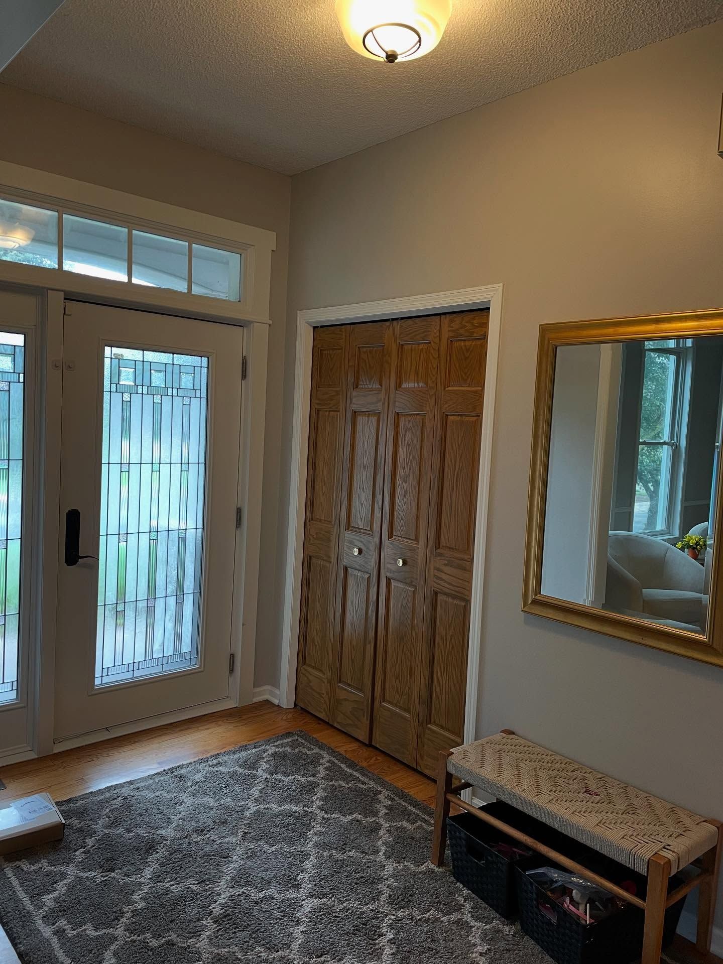 Entryway with wooden doors, a rug, and a bench with a mirror. Neutral walls and trim, glass-paned double doors to the left.