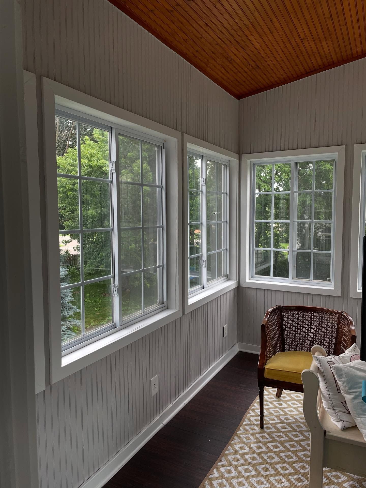 Sunroom with white paneled walls, dark wood floor, and a wood-paneled ceiling. Three multi-paned windows overlook lush greenery, and a patterned rug and chair are visible.
