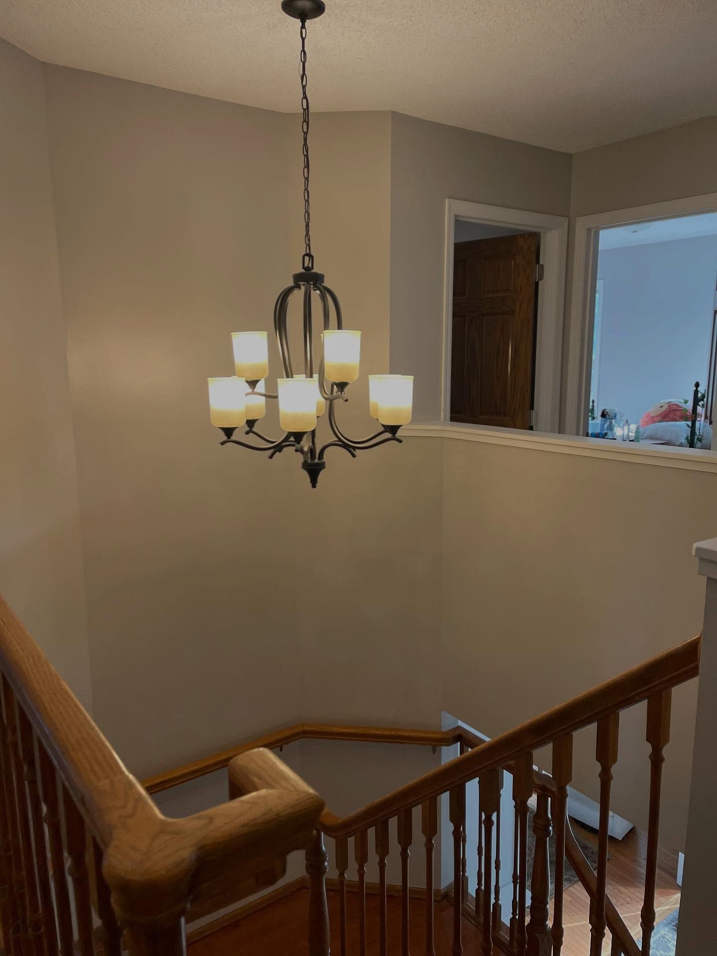 Chandelier hanging above a wooden staircase. The walls are painted beige, and a doorway is visible in the background.