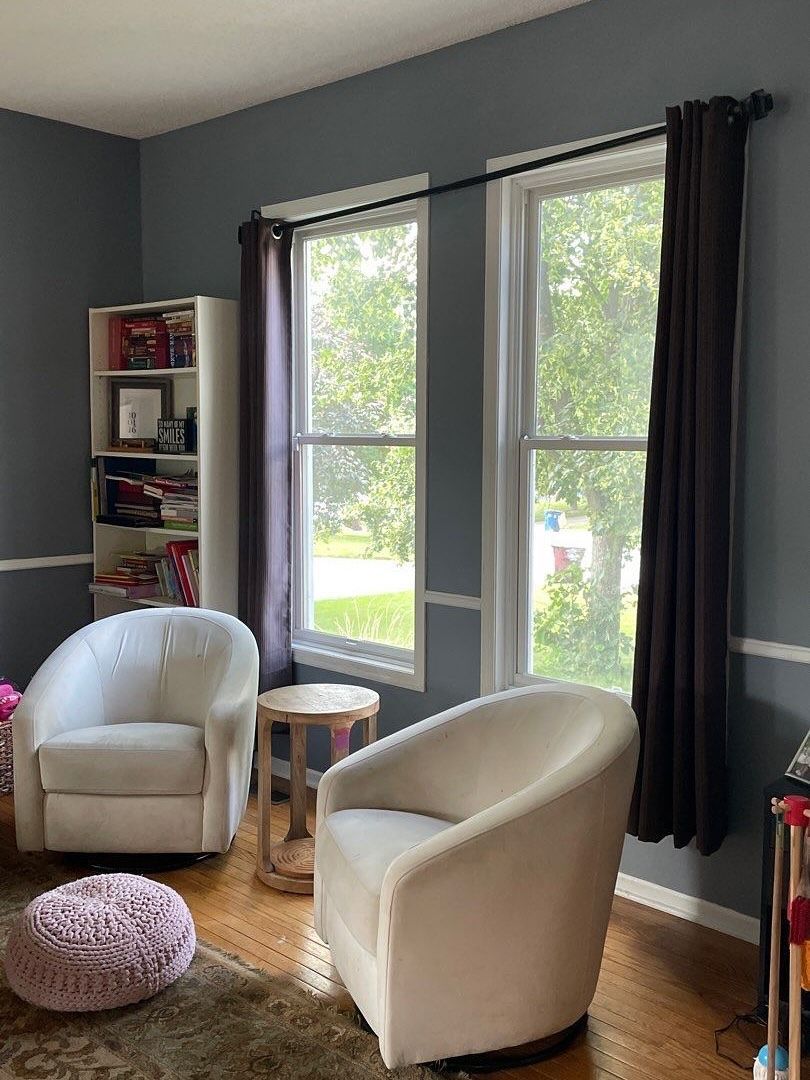 A cozy reading nook with two white swivel chairs, a small table, and dark purple curtains by a window. A bookshelf and ottoman are also present.