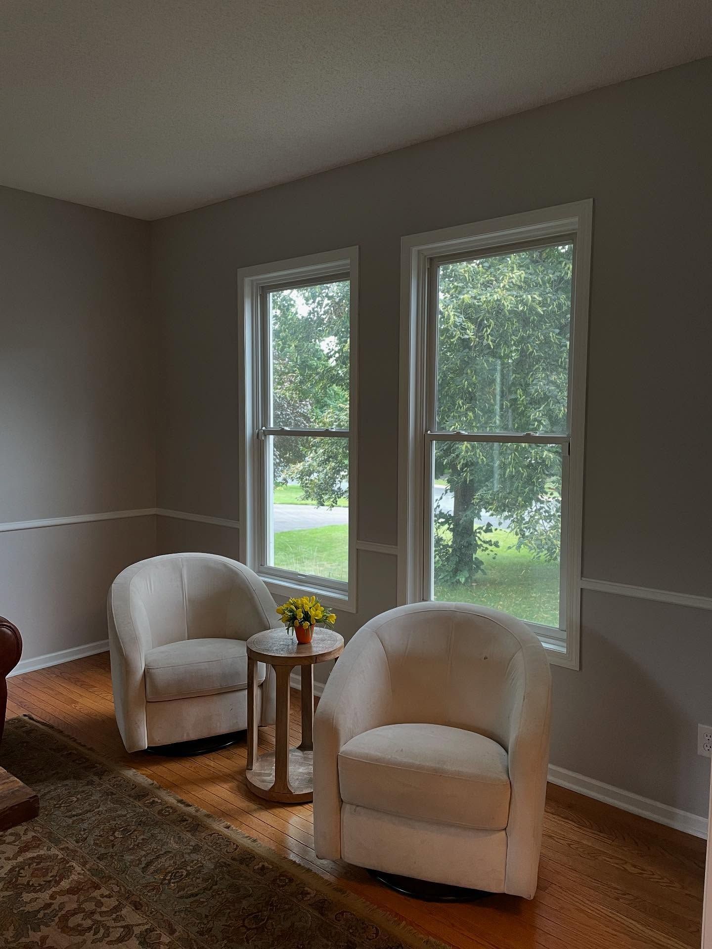 Two white armchairs face each other near two windows with a small table between them. The room has neutral-colored walls and hardwood floors.