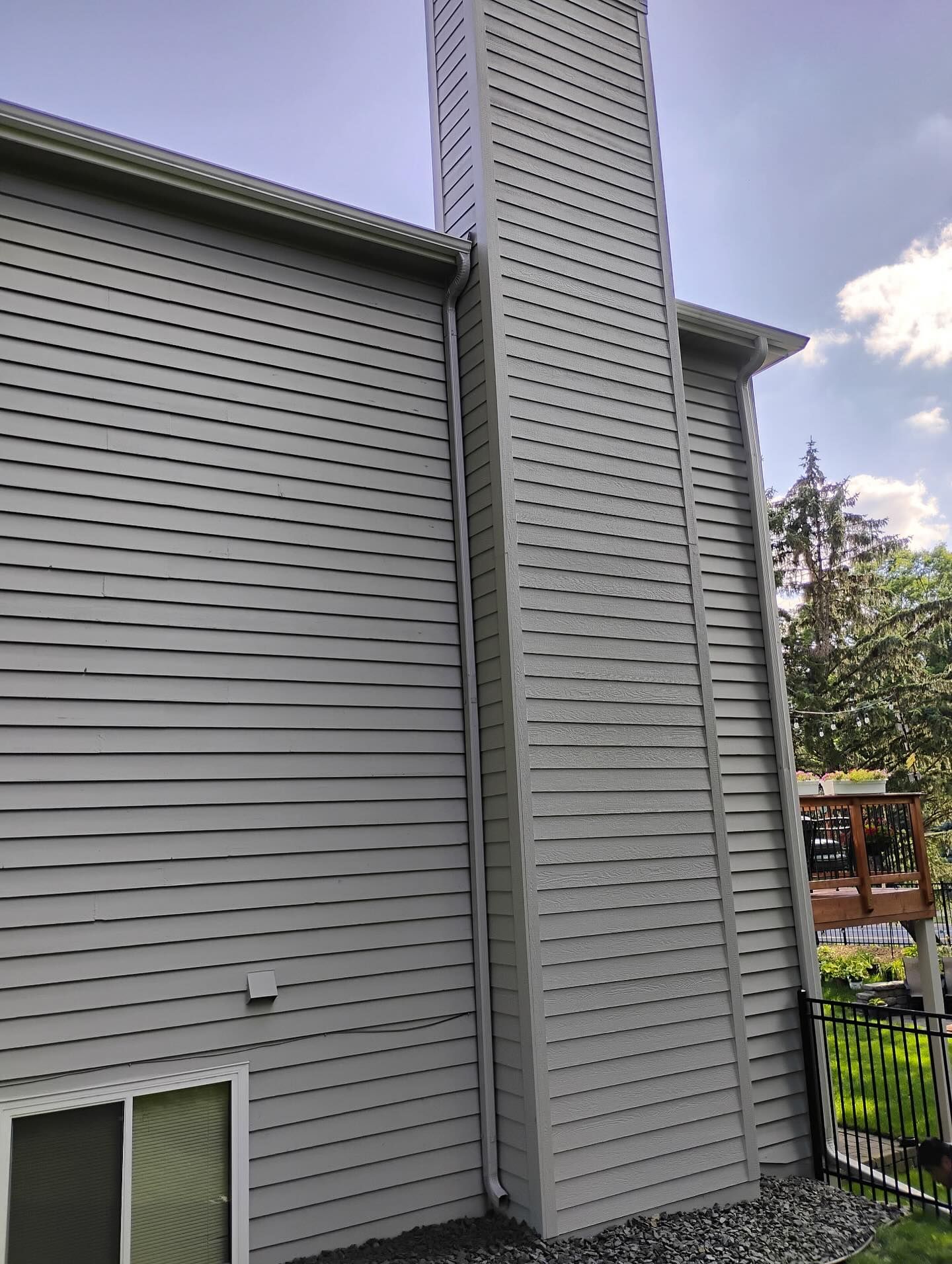 Side view of a two-story house with gray siding and a tall, matching chimney. The sky is blue with clouds.