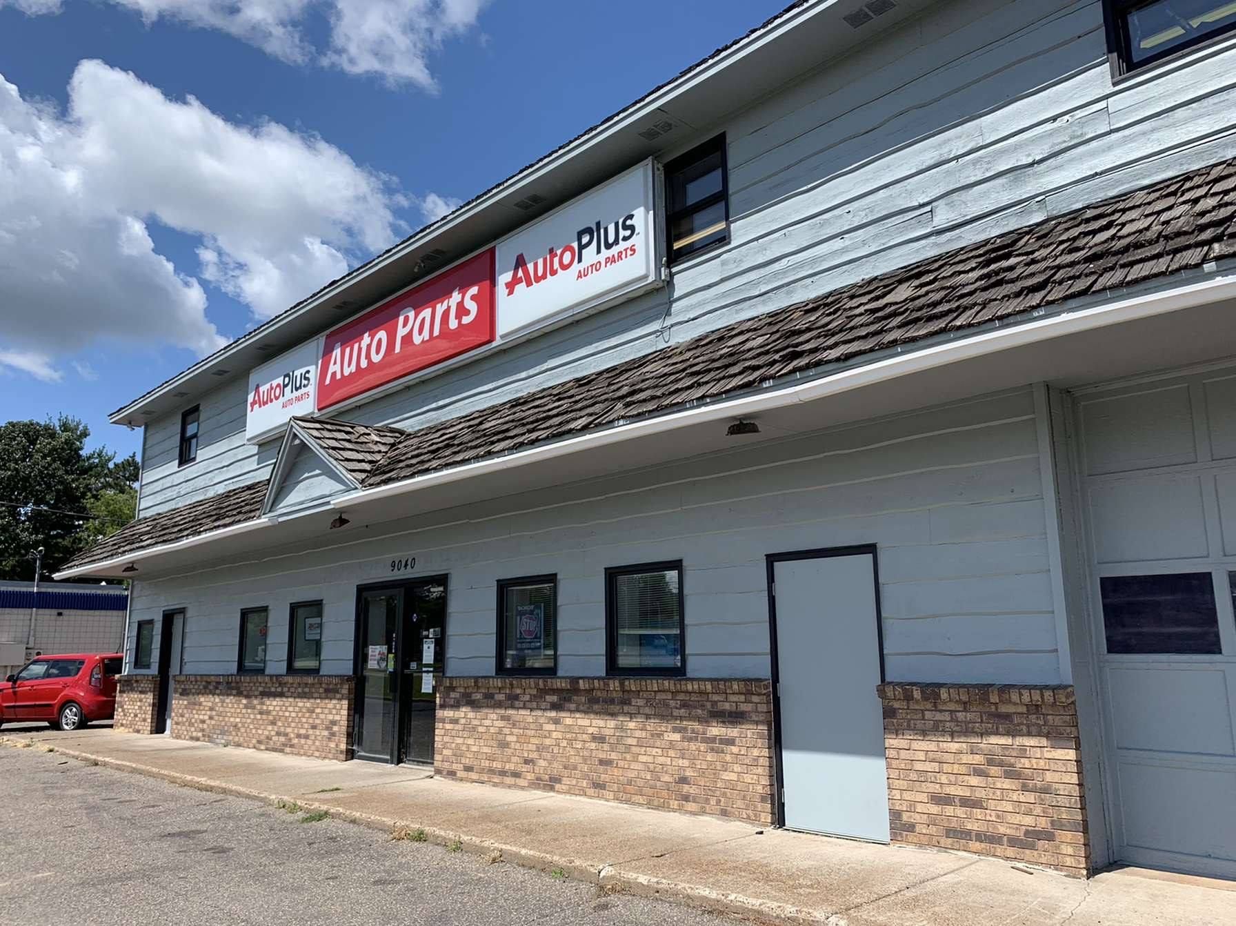Exterior view of an Auto Plus auto parts store with a red car parked nearby. The building is gray with a brick base.