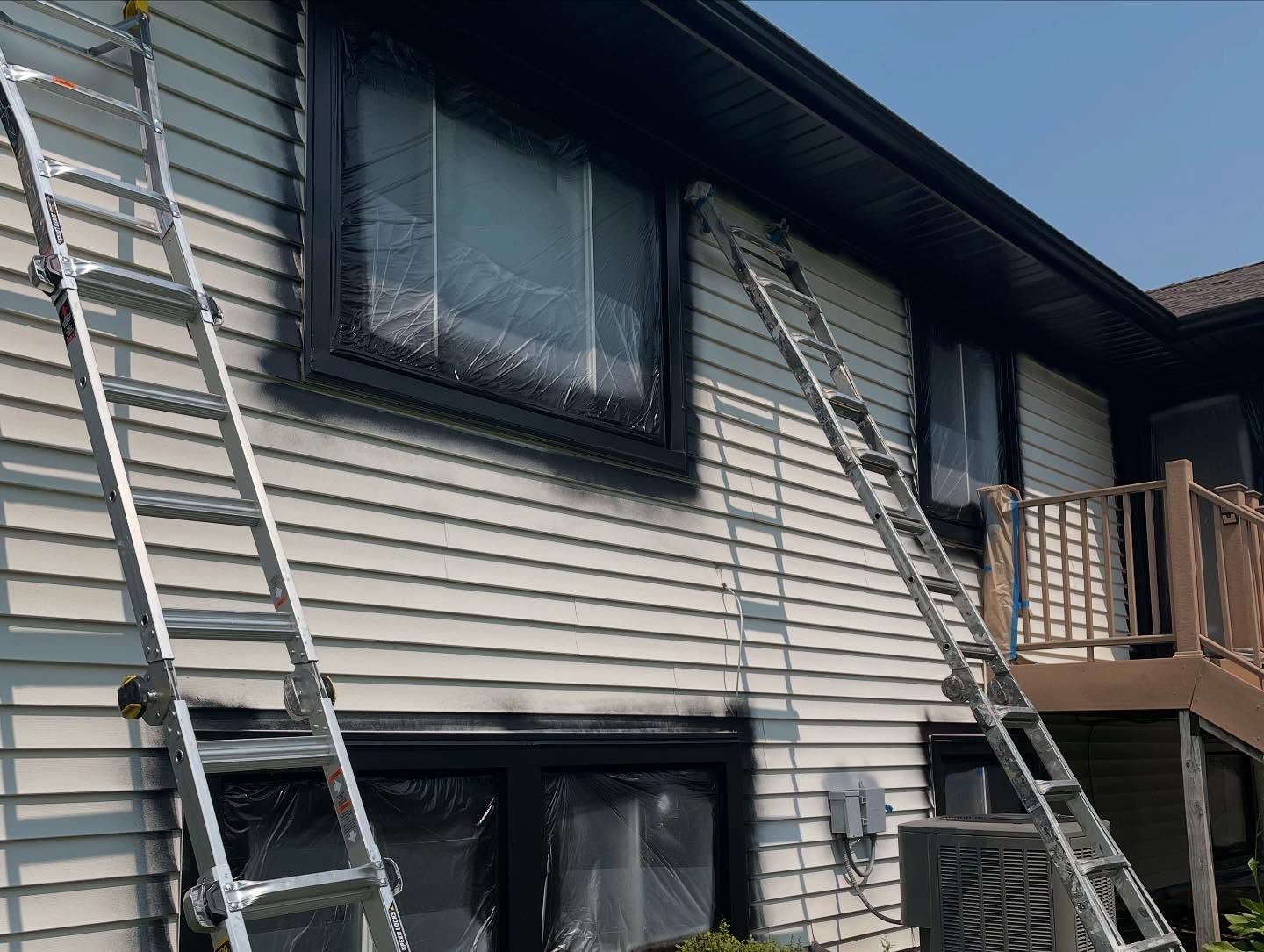 Two ladders leaning against a house with windows covered in plastic for painting; black paint around the window frames.