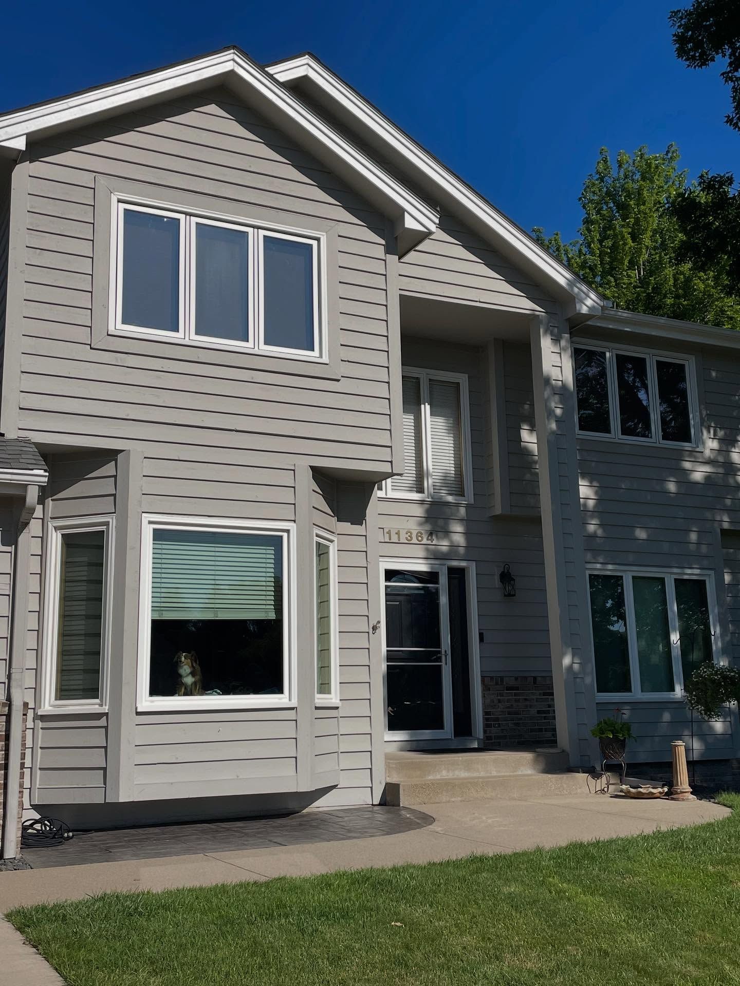 Two-story house with gray siding, white trim, and multiple windows under a clear blue sky.