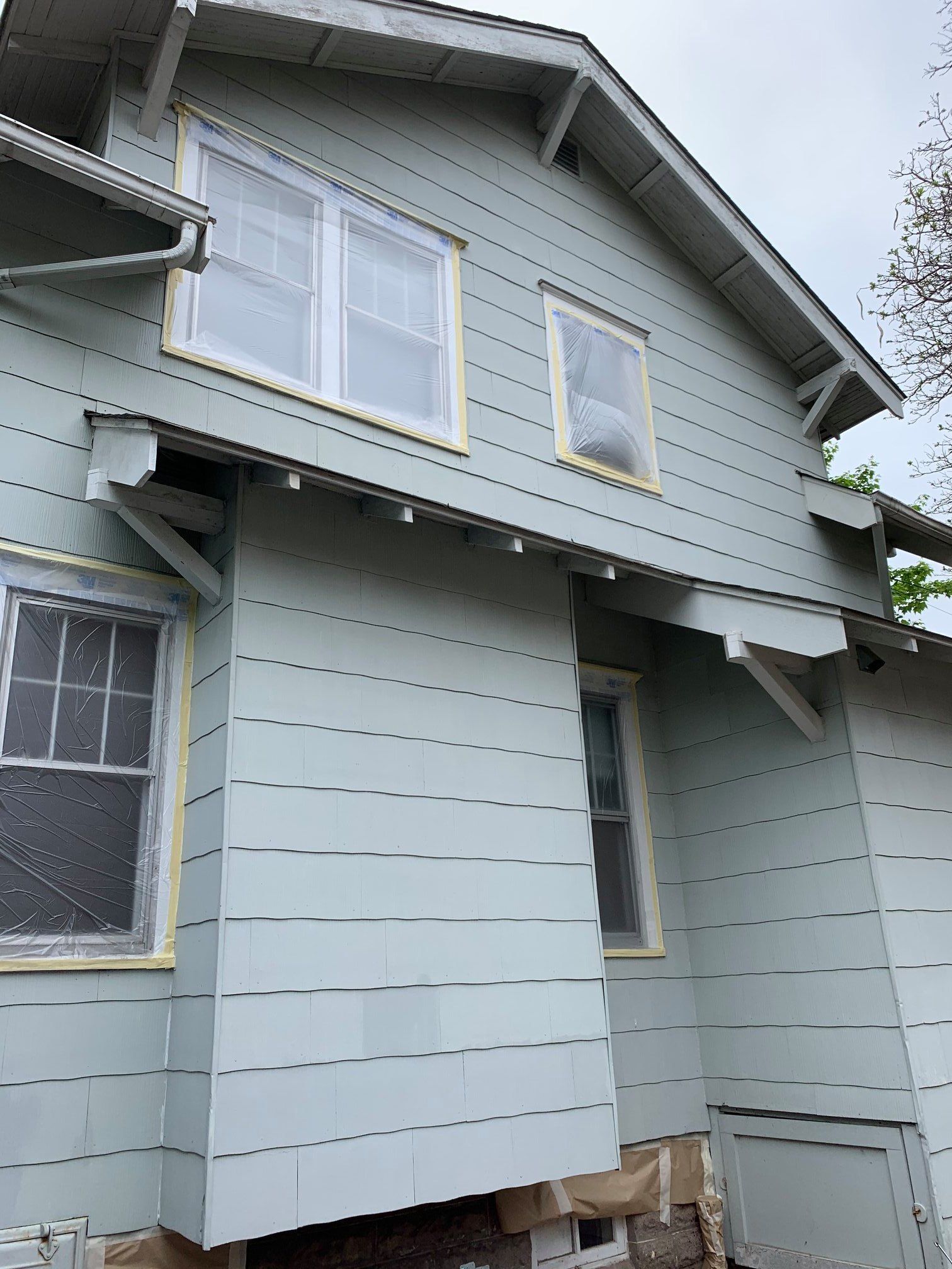 Side view of a two-story house with pale blue siding, windows taped for painting, and a partial visible foundation.