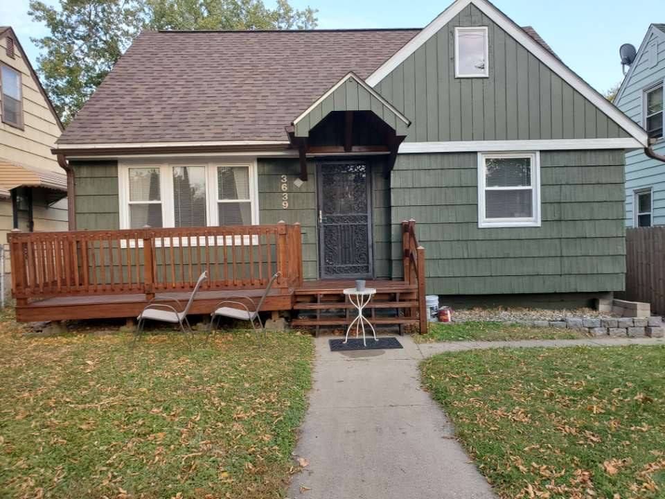 Green-sided house with a brown roof, porch, and walkway. A dark screen door and a small table sit on the porch.