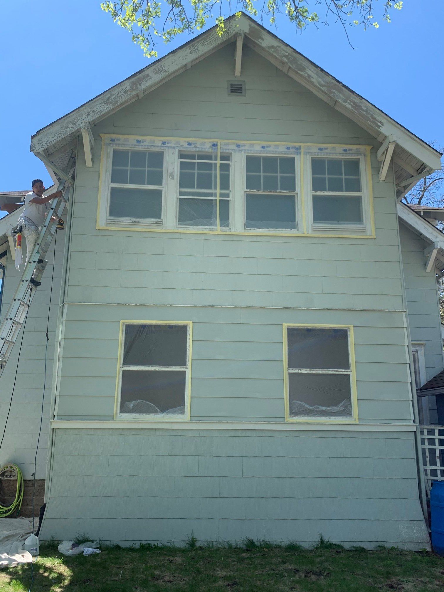 A person paints the side of a two-story house, using a ladder. The house is light green with white trim.