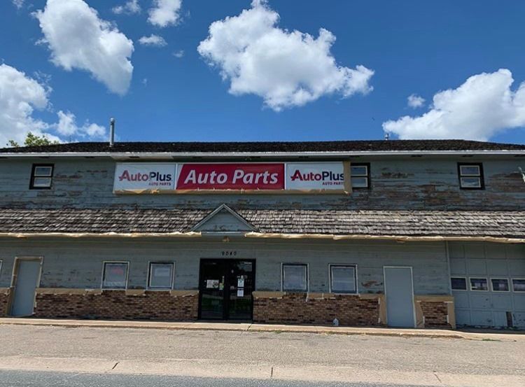 AutoPlus Auto Parts store with a weathered facade under a partly cloudy sky. The building has a brick base and a central entrance.