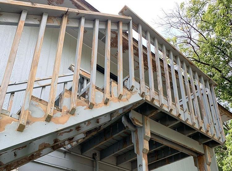 A weathered wooden deck with peeling paint. Railings and stairs are visible, showing signs of age and disrepair.