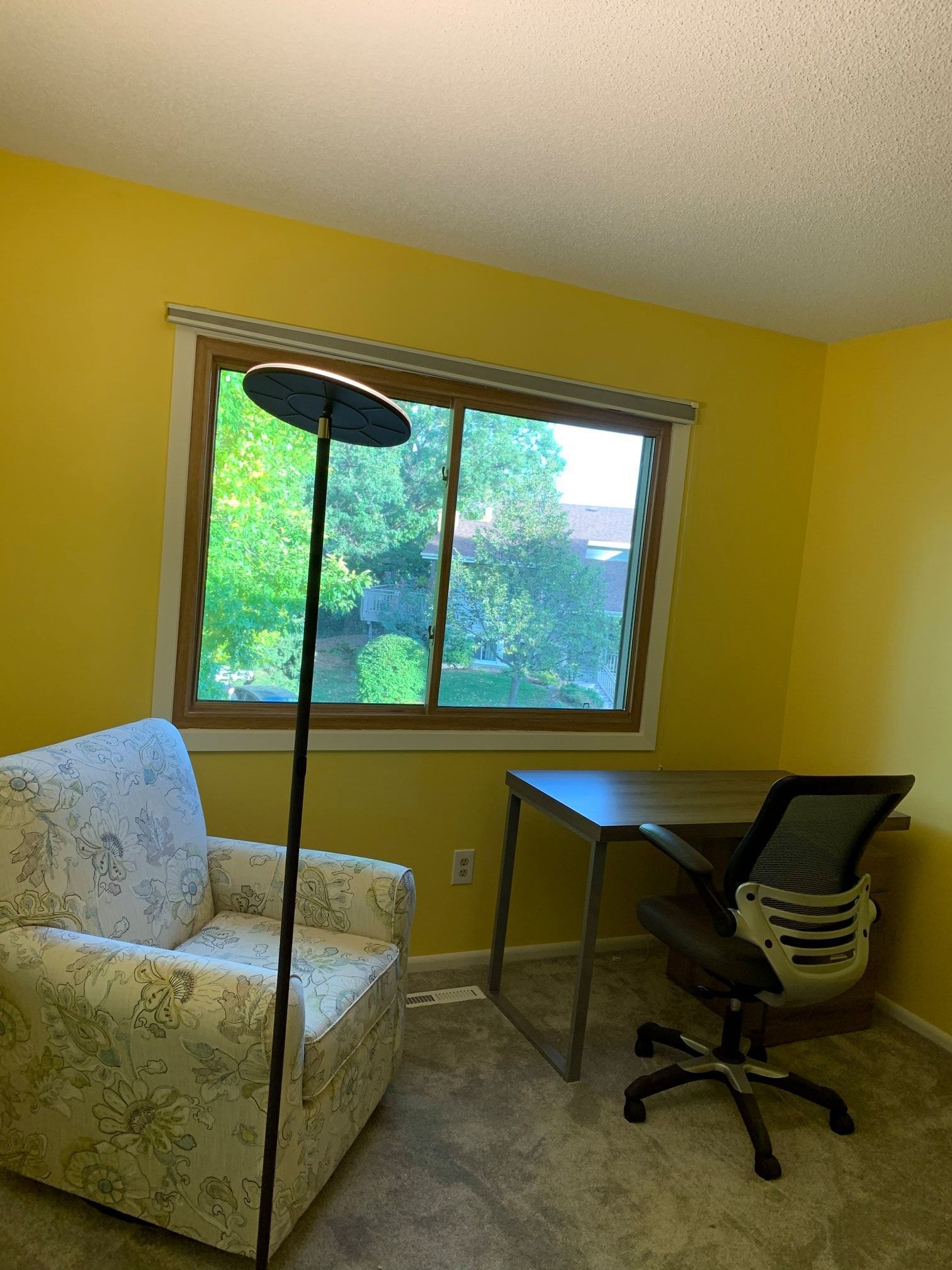 A yellow-walled room with a floral armchair, a desk with an office chair, and a tall lamp near a window.