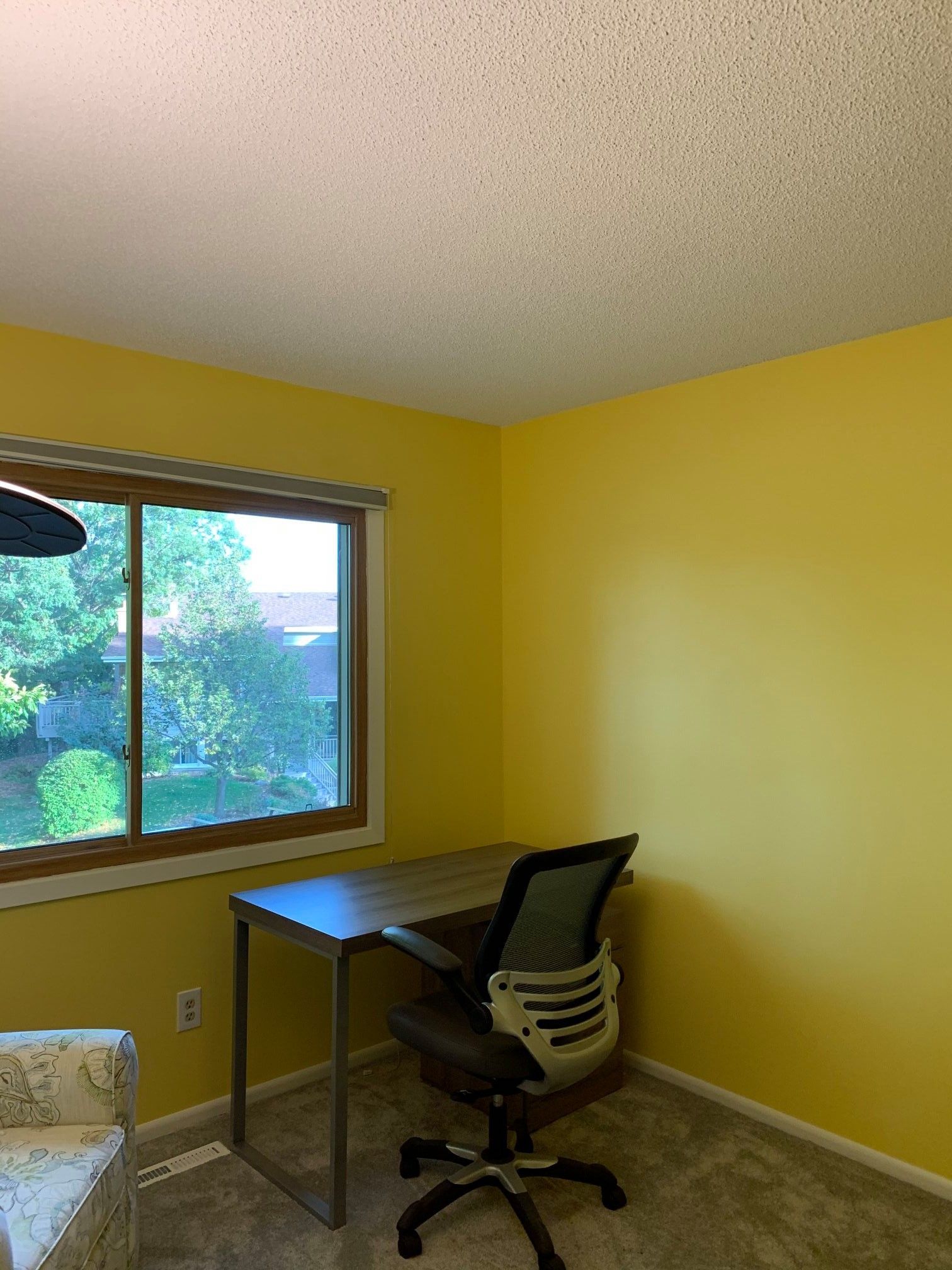 Yellow-walled home office with a desk, chair, and window overlooking a green landscape. A corner chair is visible.