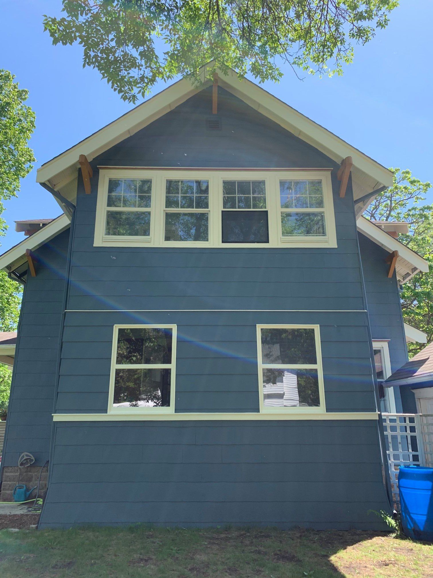 Two-story blue house with light yellow window frames and wooden accents. Sunlight shines on the house; trees in the background.