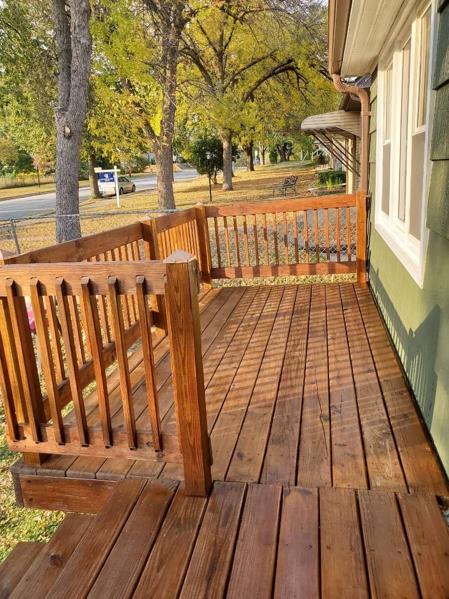 Wooden porch with a railing, next to a green house. Trees and a street are visible in the background.