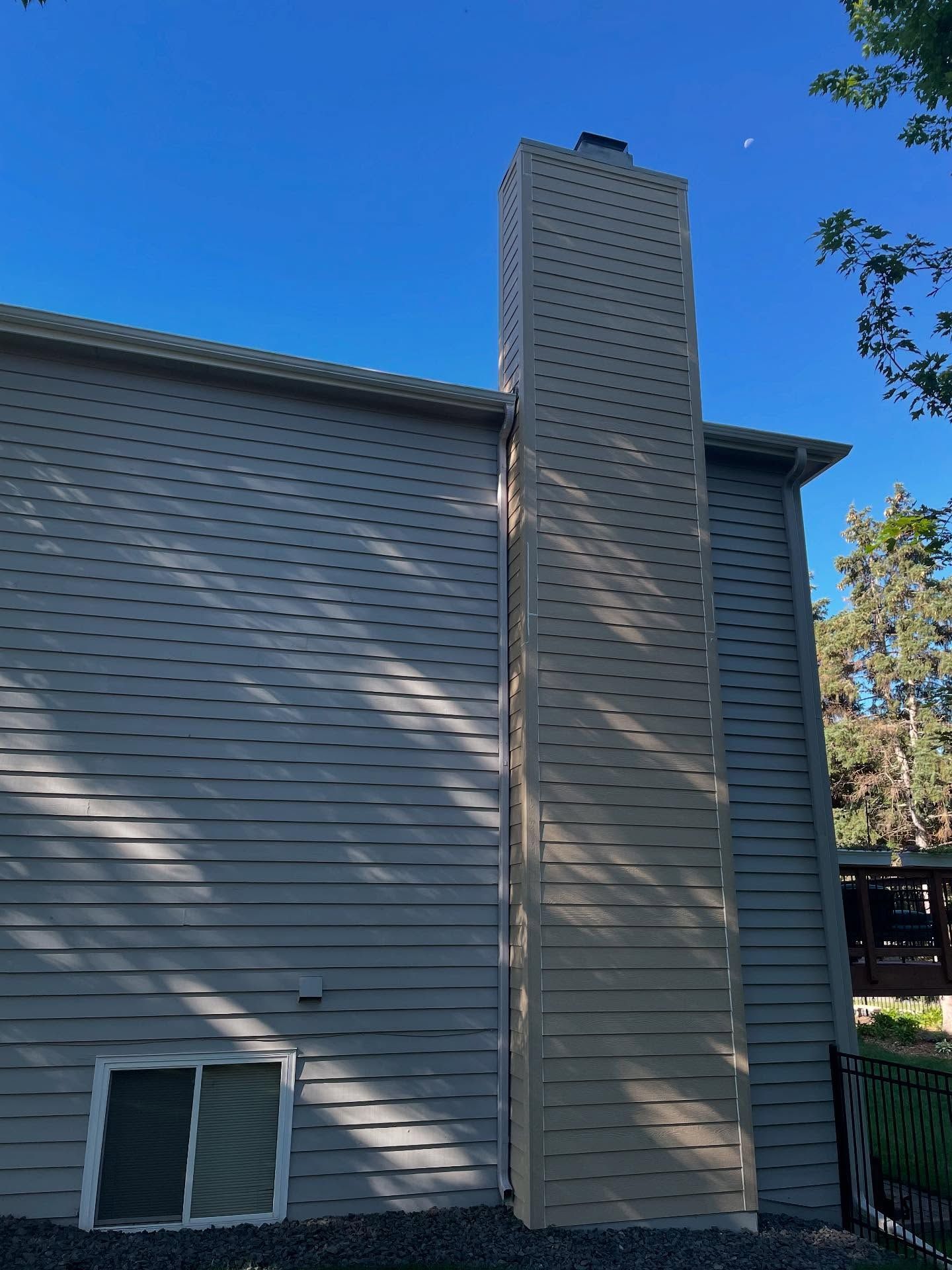 Tall, beige chimney on the side of a two-story house. The sky is blue, and the house has gray siding.