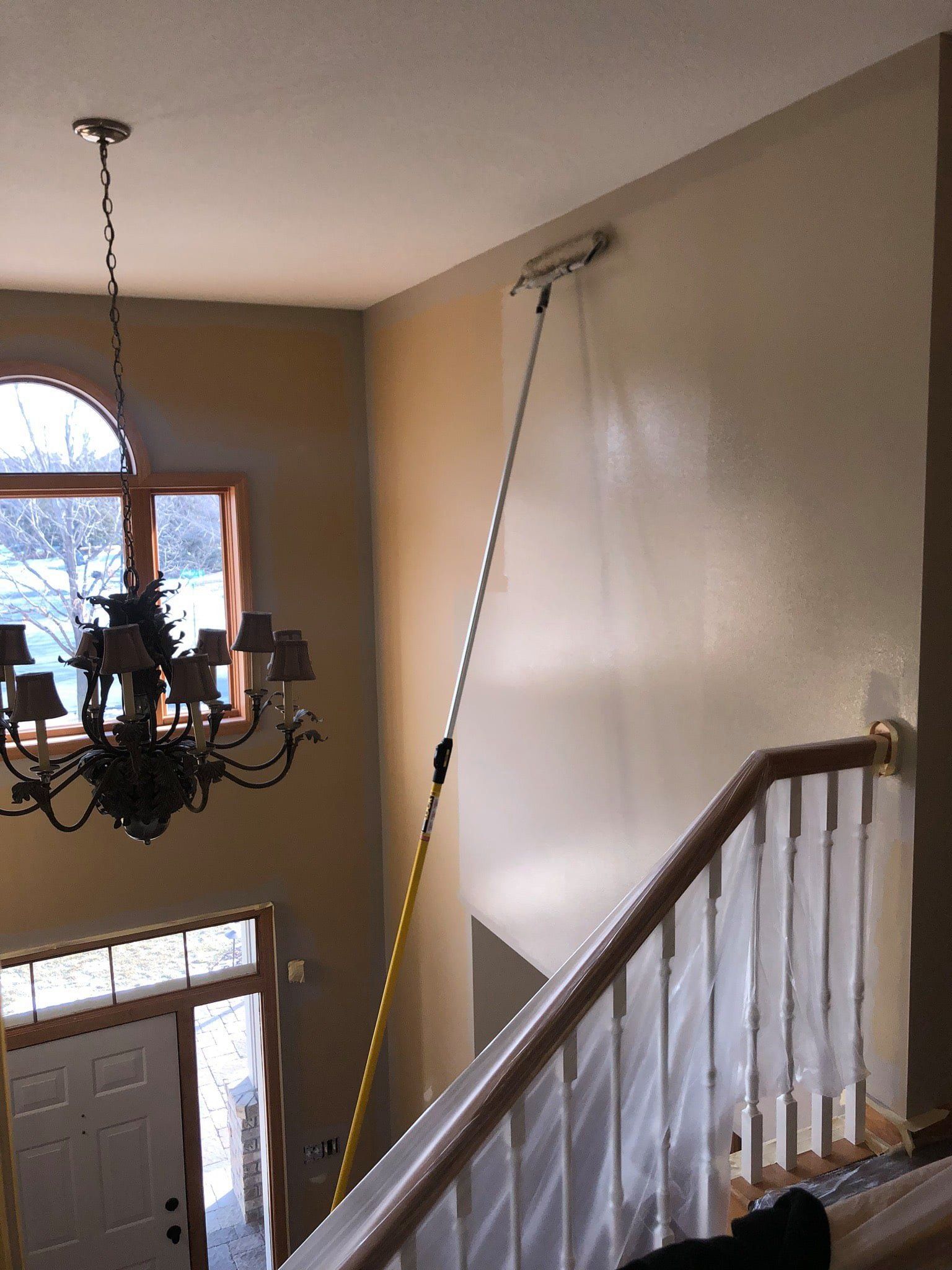 Person painting a tall beige wall with an extended roller. A chandelier hangs near a window and a staircase is covered in plastic.