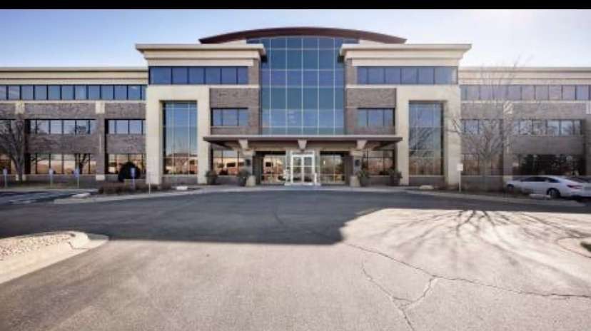 Modern office building with glass and stone facade, gray and beige tones, clear sky, and parking lot in front.